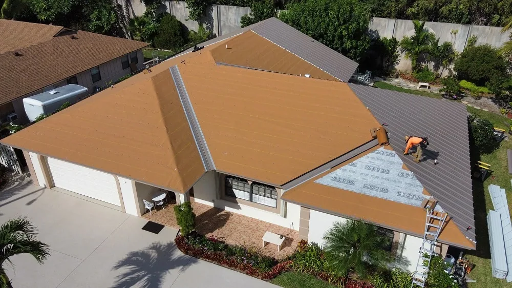 Aerial view of a single-story home with tan underlayment and gray metal panels being installed by a roofer near a ladder