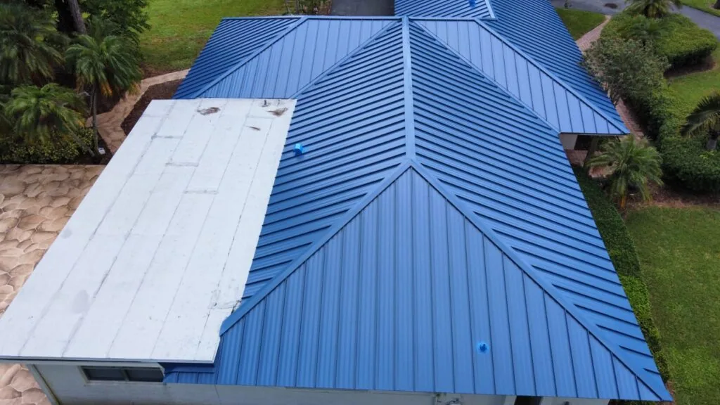 Aerial view of a blue standing seam metal hip roof next to a white flat roof section on a single-story home with landscaping