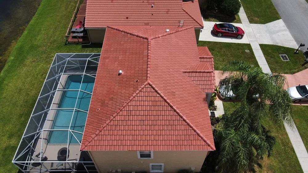 Aerial view of a red barrel tile hip roof on a stucco home with a screened pool enclosure, palm tree, and lawn beside a lake