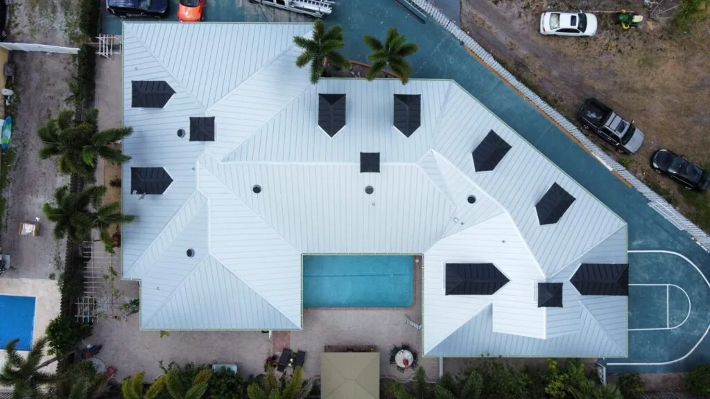 Top-down aerial view of a white standing seam metal roof with dark dormer roofs, a rectangular pool, and a paved patio