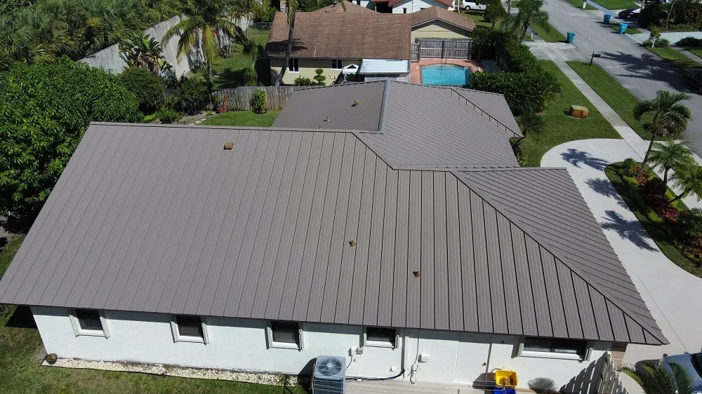Aerial view of a taupe standing seam metal roof on a white stucco single-story house with a neighboring pool and palm trees