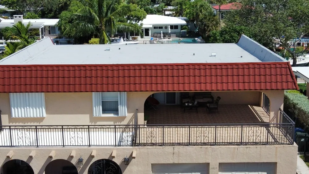 Two-story stucco home with a white flat roof, red barrel tile roof edge, covered balcony with iron railing, and homes behind