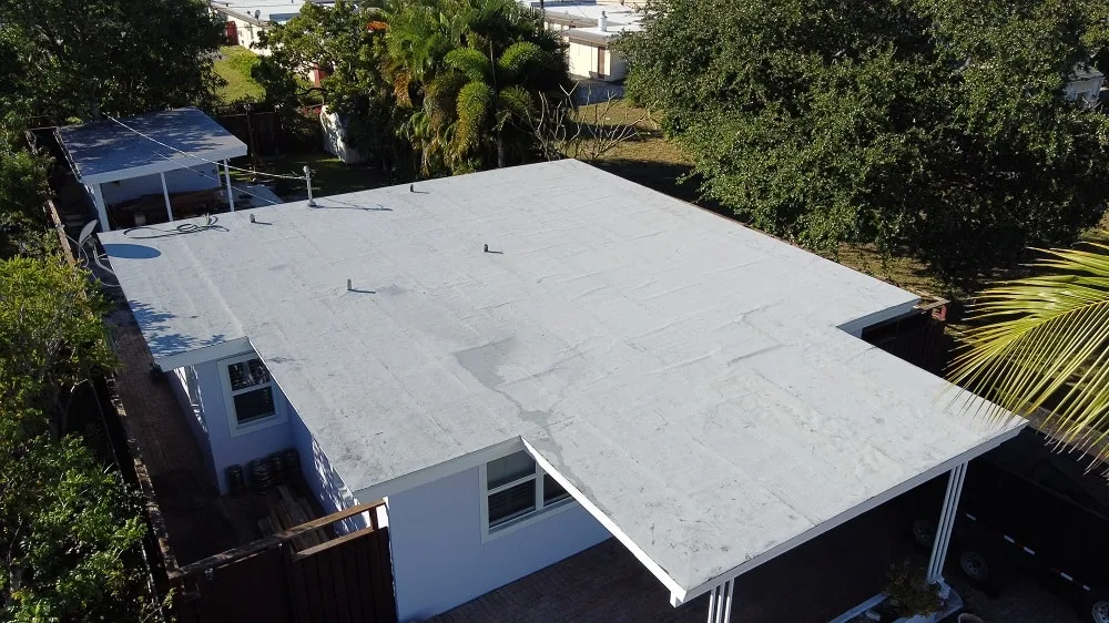 Aerial view of a white flat roof with vent pipes on a single-story home with a carport, fenced yard, and palm trees