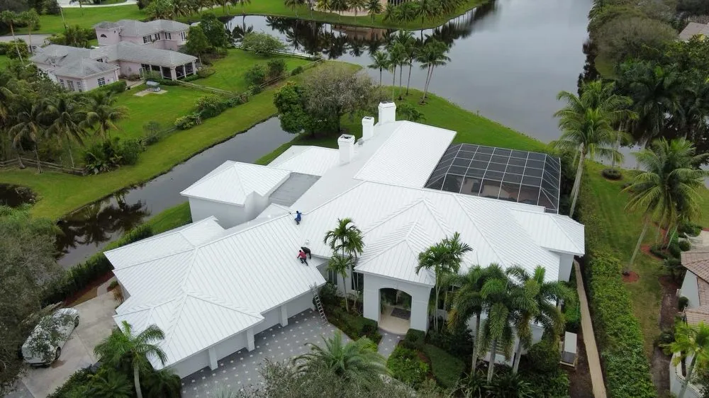 Aerial view of workers on a white standing seam metal roof of a large home beside a canal with a screened pool