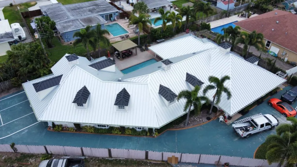 Aerial view of a large home with a white standing seam metal roof, dark gray dormer caps, a pool, palm trees and a work truck