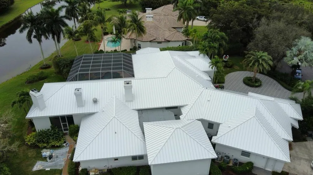 Aerial view of a white standing seam metal roof with several hips and chimneys on a large home beside a lake and screened